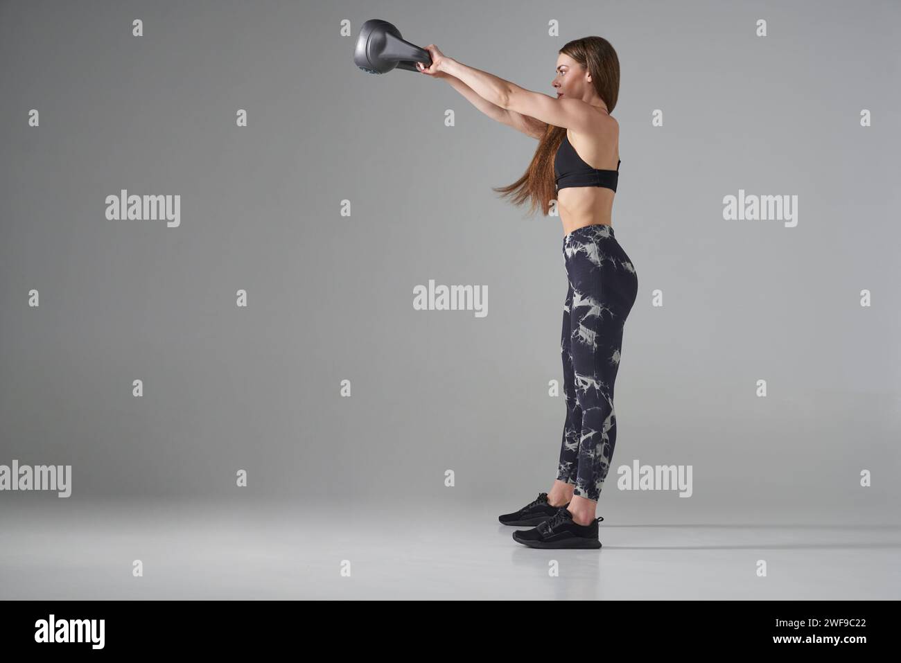 Motivated young sportswoman in legging, doing weight exercises with kettle bell. Side view of slim, muscular long-haired female lifting kettle bell an Stock Photo