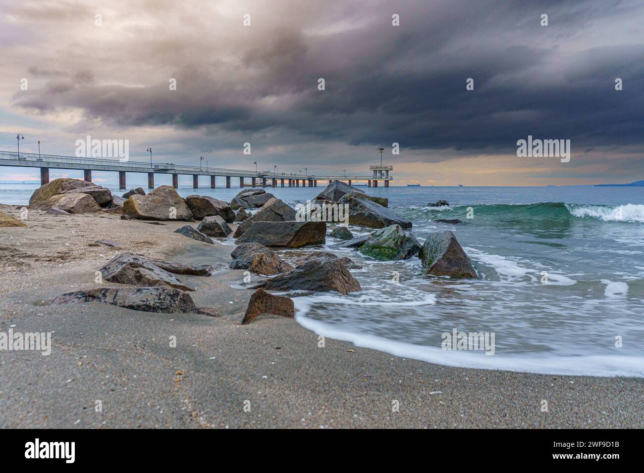 A pier extending over calm waters, adjacent to a sandy beach, beneath overcast skies Stock Photo