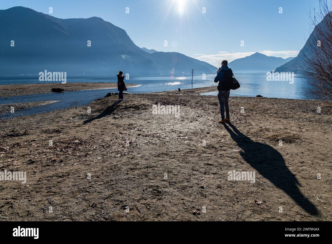 A couple playing with their dog and admiring the scenic view of Lake Lugano in the Alps on a sandy beach in Lugano, Switzerland Stock Photo
