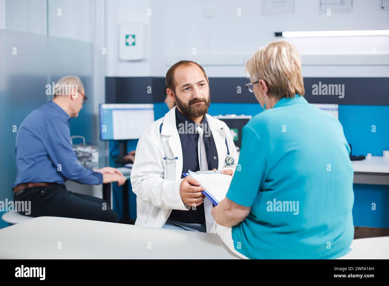 Senior woman filling checkup paperwork and discussing treatment with male doctor. Physician requests patient signature on paperwork in order to treat orthopedic injury with physiotherapy. Stock Photo