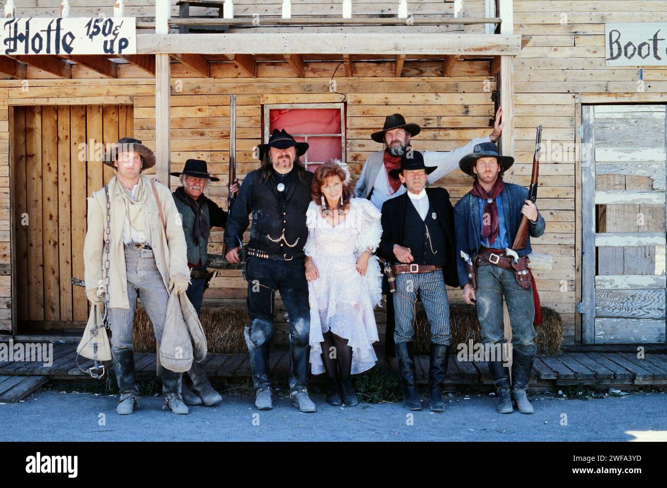 A group of seven people stands in a line, posing in period-specific costumes representative of the American Wild West era, in front of a rustic saloon Stock Photo