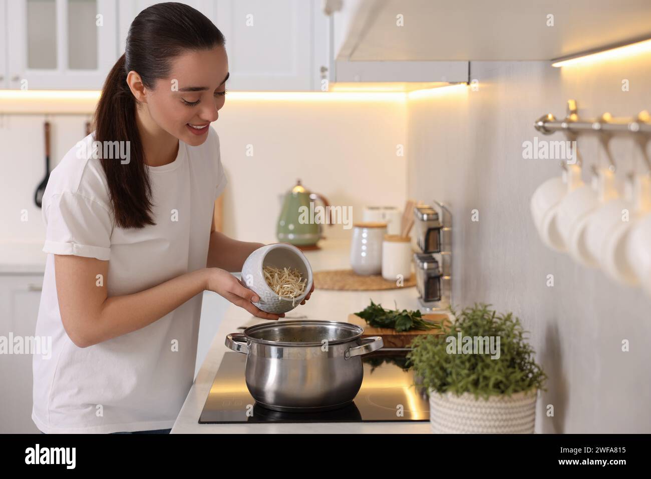 Smiling woman adding noodles into pot with soup in kitchen Stock Photo