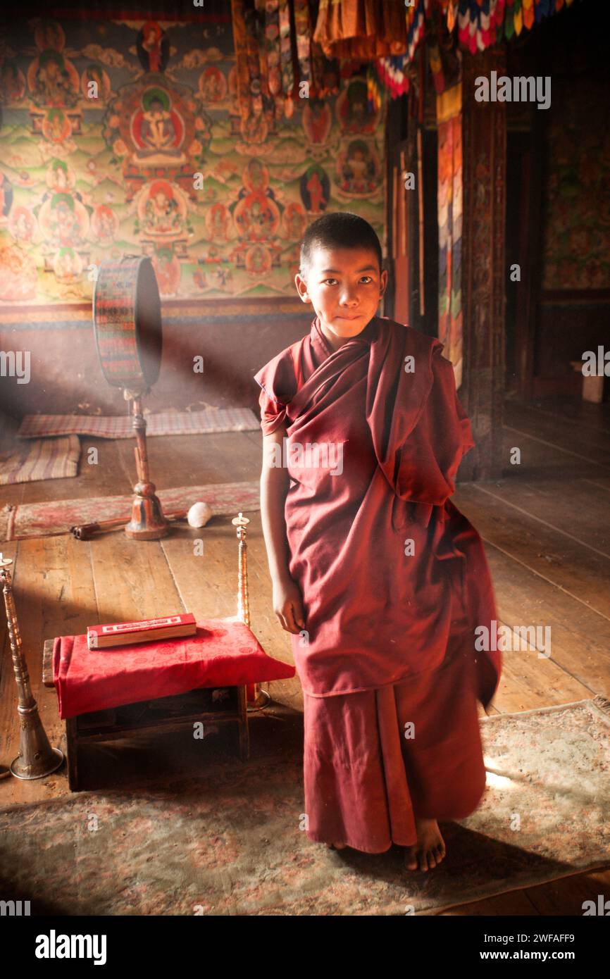 A young barefoot monk in a red robe stands on a weathered rug over a wooden floor inside the ancient Buddhist temple where he resides in Ura village Stock Photo