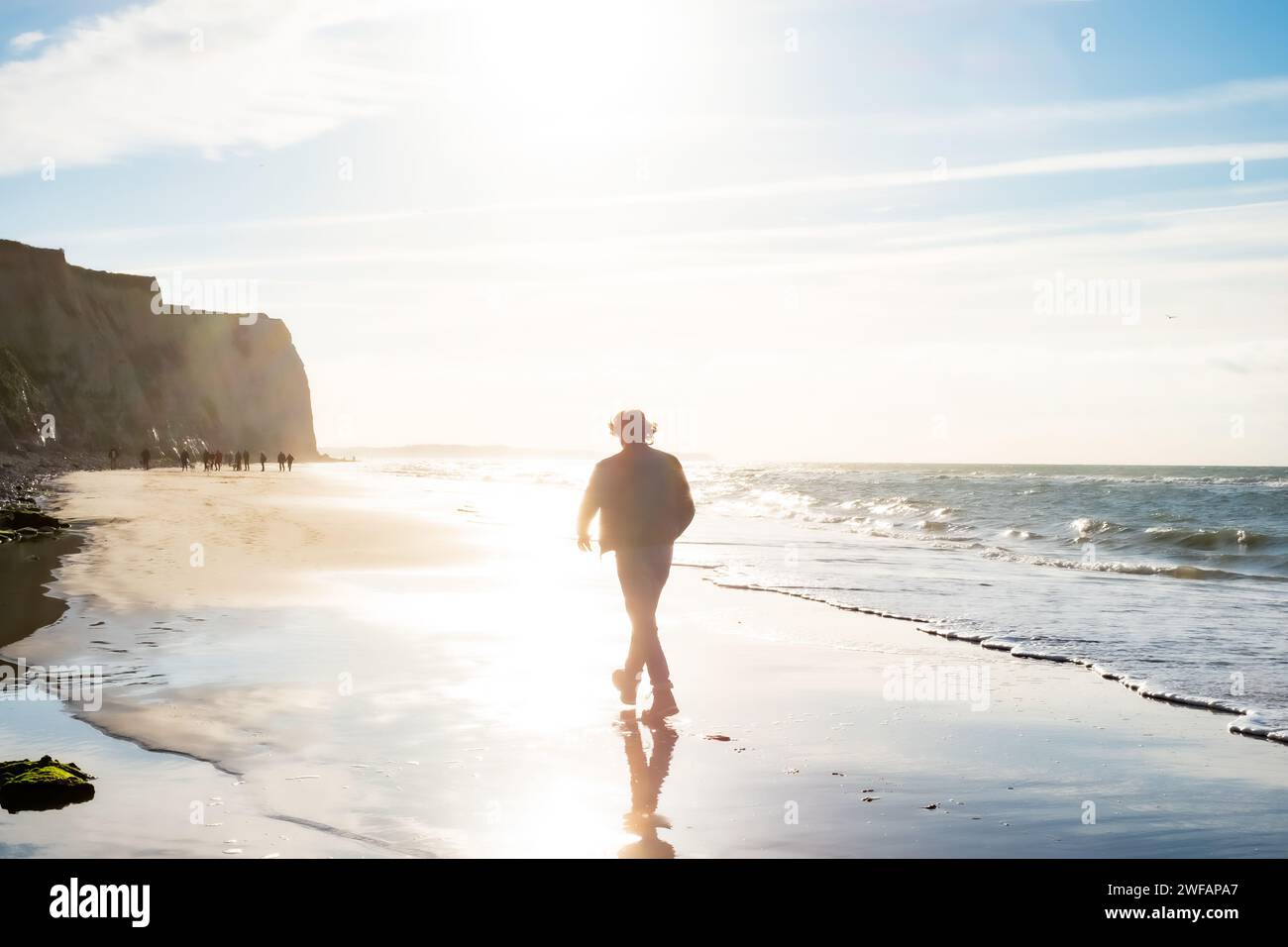 The image presents a lone individual embarking on a leisurely walk along the shoreline, with the dawn sun casting a diffuse light across the scene. This time, the person appears closer and more prominent, suggesting a narrative of engagement with the environment. The morning light softens the outlines of the cliffs and the figure, creating a dreamlike quality. Small waves break gently on the shore, while the wet sand reflects the figure and the glittering sunlight, enhancing the sense of early morning tranquility. Coastal Journey at Dawn. High quality photo Stock Photo