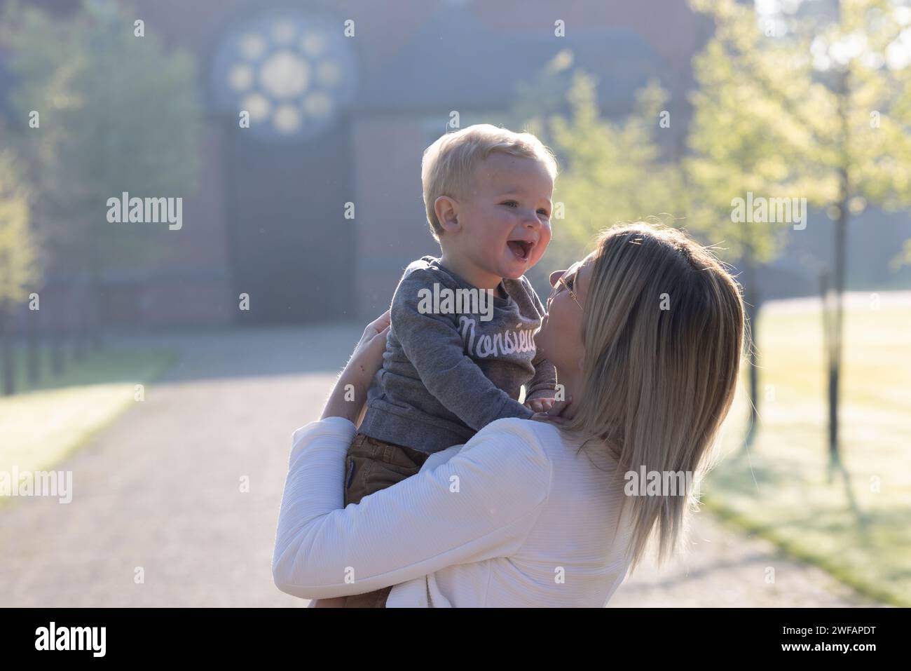 This delightful capture features a young Caucasian woman lifting a toddler into the air, both basking in the brilliance of a joyful morning. As the toddler laughs heartily, his eyes squinting in merriment, the woman gazes at him with an expression of love and happiness. The ambient light of the early day surrounds them, enhancing the exuberant and loving energy of their interaction. Gleeful Mornings: A Symphony of Laughter. High quality photo Stock Photo