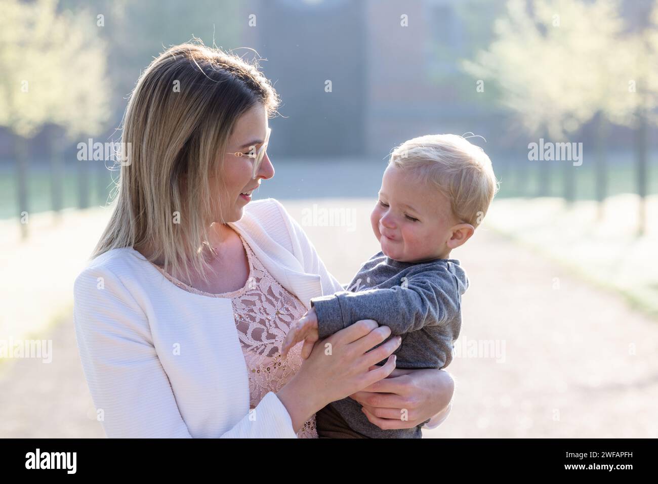 The image radiates with the warmth of a maternal bond as a young Caucasian woman holds a toddler, their gazes locked in a loving exchange. The softness in the child's eyes and the gentle smile on the woman's lips speak volumes about their special connection. The gentle morning light washes over them, enhancing the tenderness of the moment, set against the tranquil backdrop of a sunlit park. Intimate Moments of Maternal Bliss. High quality photo Stock Photo