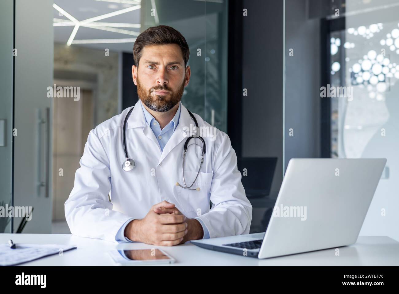 Portrait of a young male doctor sitting in a slaughterhouse office at a desk in a white coat, looking seriously and focused into the camera. Stock Photo