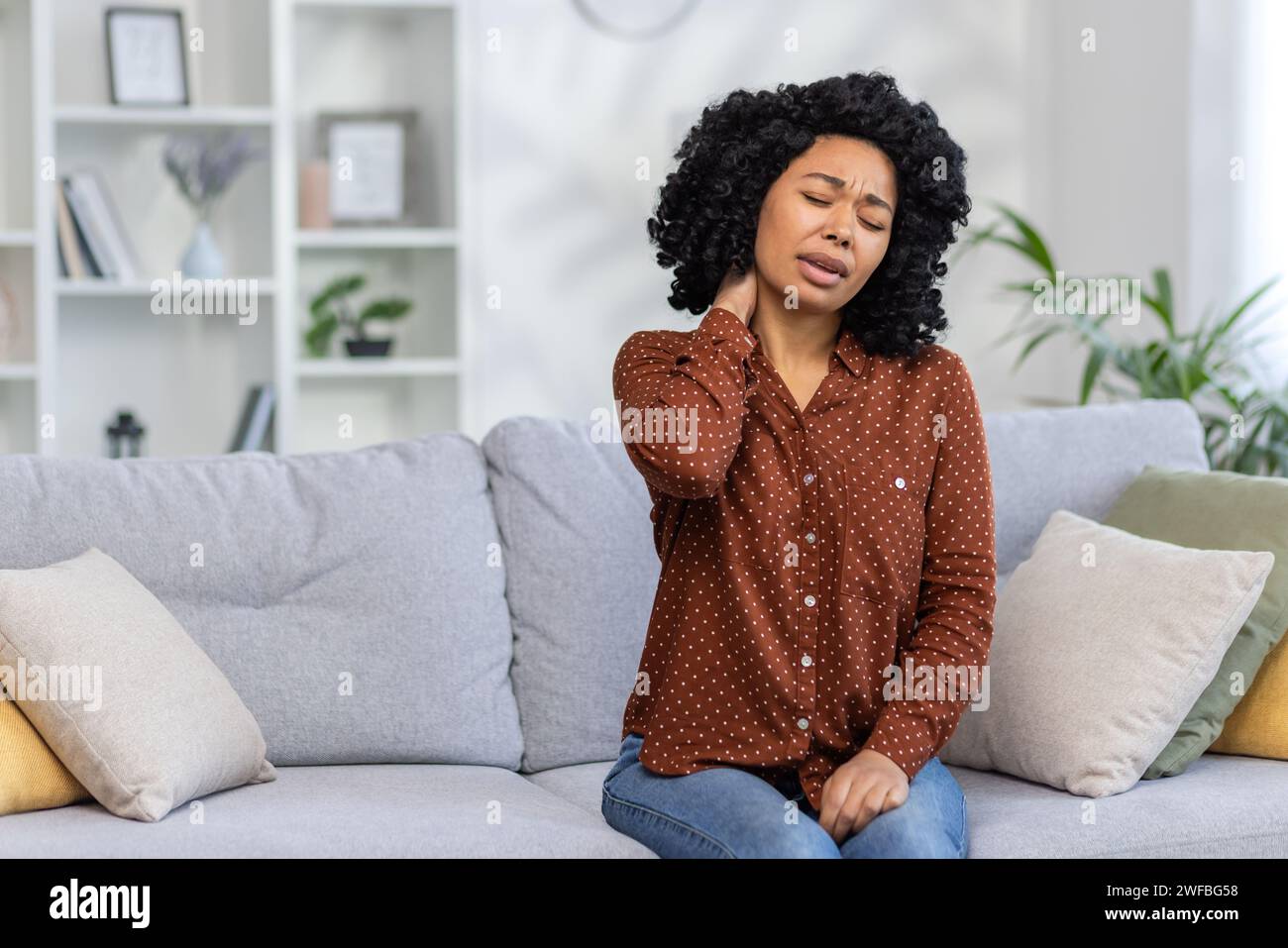 African American young woman sitting at home on sofa tired, closing eyes resting and doing neck massage, feeling pain and tension. Stock Photo