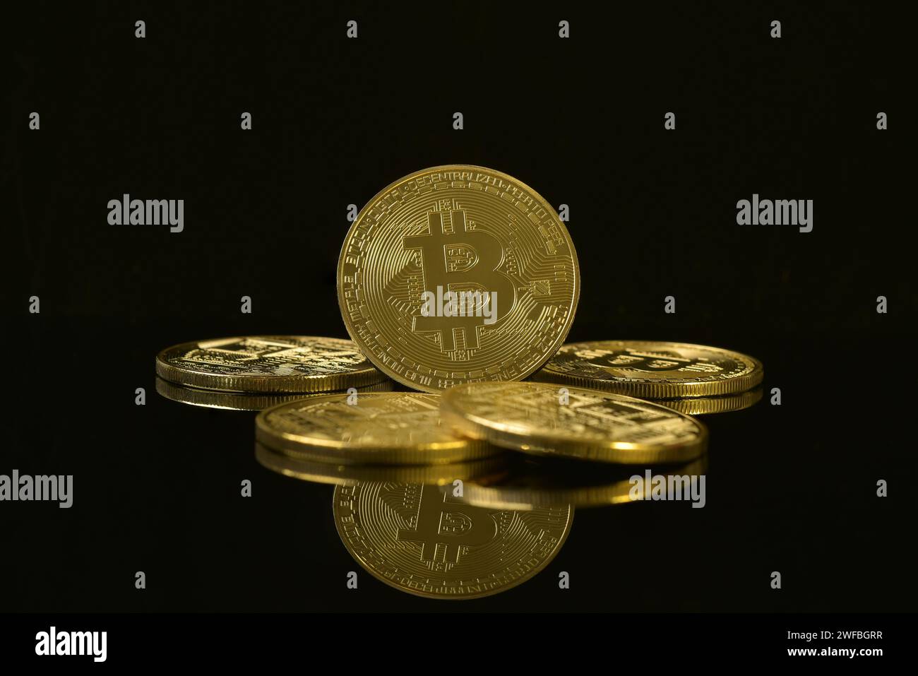 Photograph of golden Bitcoin isolated on black reflective background, surrounded by pile of coins. Cryptocurrency. Decentralised digital currency. Stock Photo