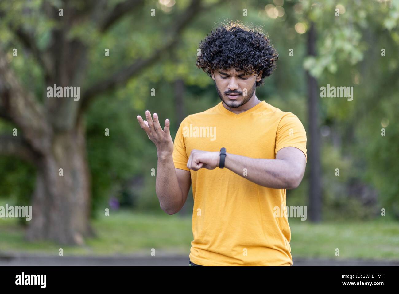 Focused young man with curly hair checking smartwatch during outdoor workout in park, wearing casual yellow t-shirt, blurred greenery. Stock Photo
