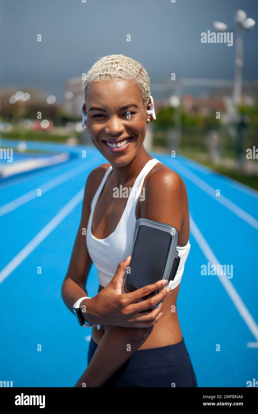 Smiling athletic sportswoman wears earphones and touch smartphone in her armband. Tracks her running sports training and jogging. African American wom Stock Photo