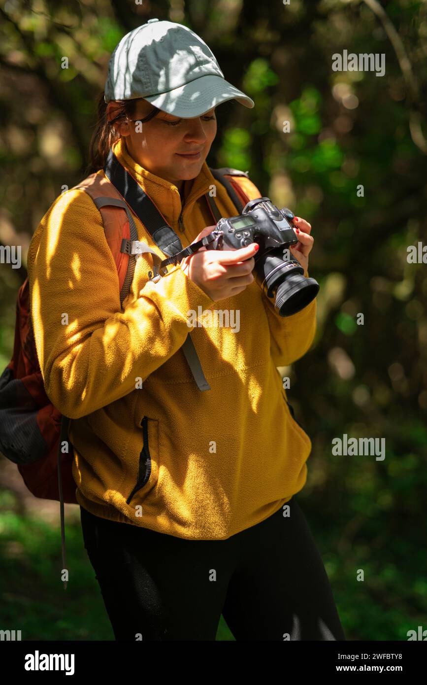 Beautiful Latin American woman walking with a red backpack and blue cap reviewing the photos on her camera in the middle of a forest during a sunny da Stock Photo