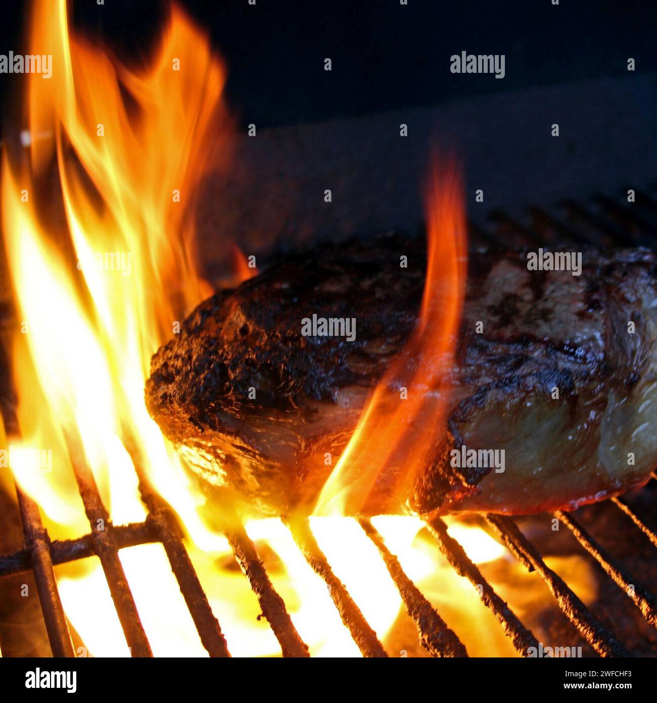 Rib eye on a grill, surrounded by flames. Searing the steak before cooking at a reduced temperature. Extra thick cut of ribeye steak on a grill top. Stock Photo