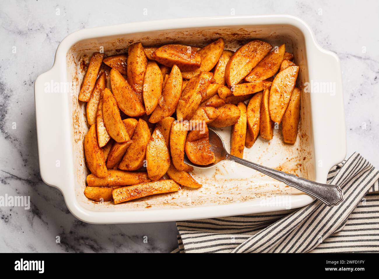 Baked apple slices with cinnamon and maple syrup in a white baking pan, top view. Stock Photo
