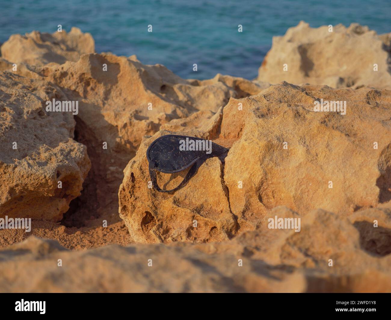 Dirty black sleeping mask covered in dust and sand lays lost and forgotten on a big porous rock on the shore of the cyan Mediterranean sea in Cyprus. Stock Photo