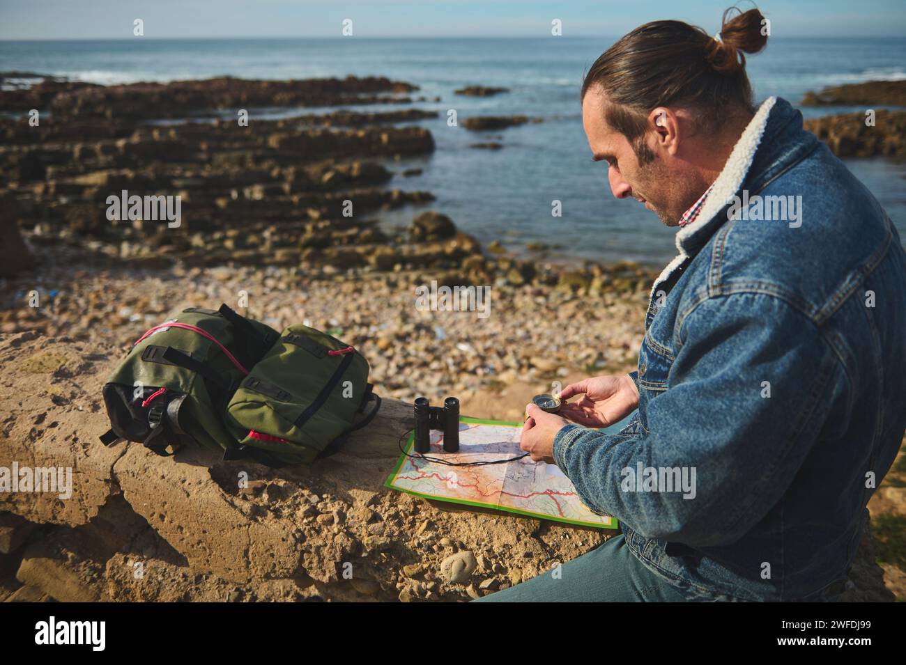 Handsome relaxed young adult man, hiker tourist sitting on he cliff and orienteering in the nature with compass in the hand, reading map and searching Stock Photo