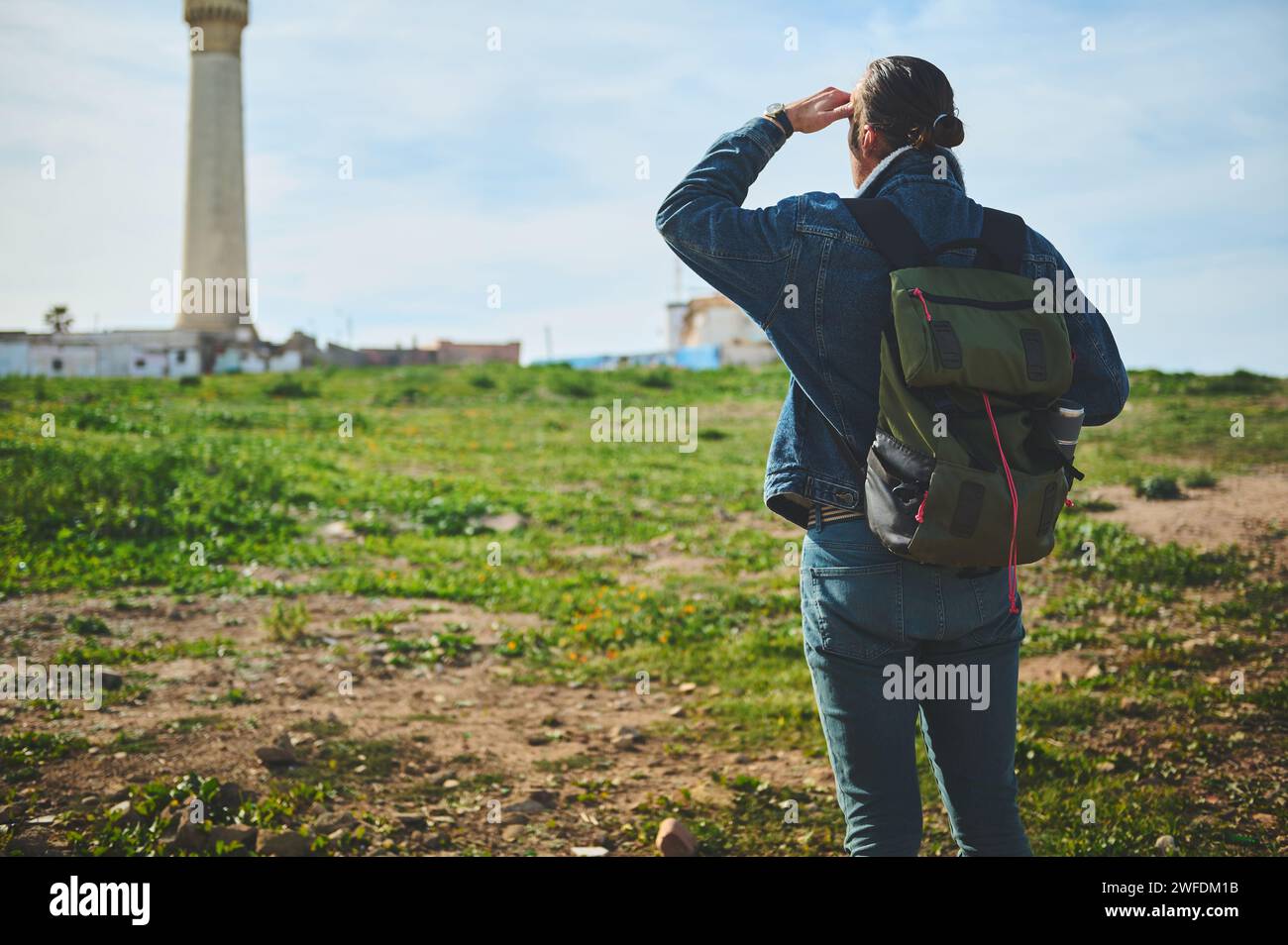 Rear view of a traveler hiker man with backpack, standing back to the camera and looking into the distance, standing over a lighthouse background on a Stock Photo