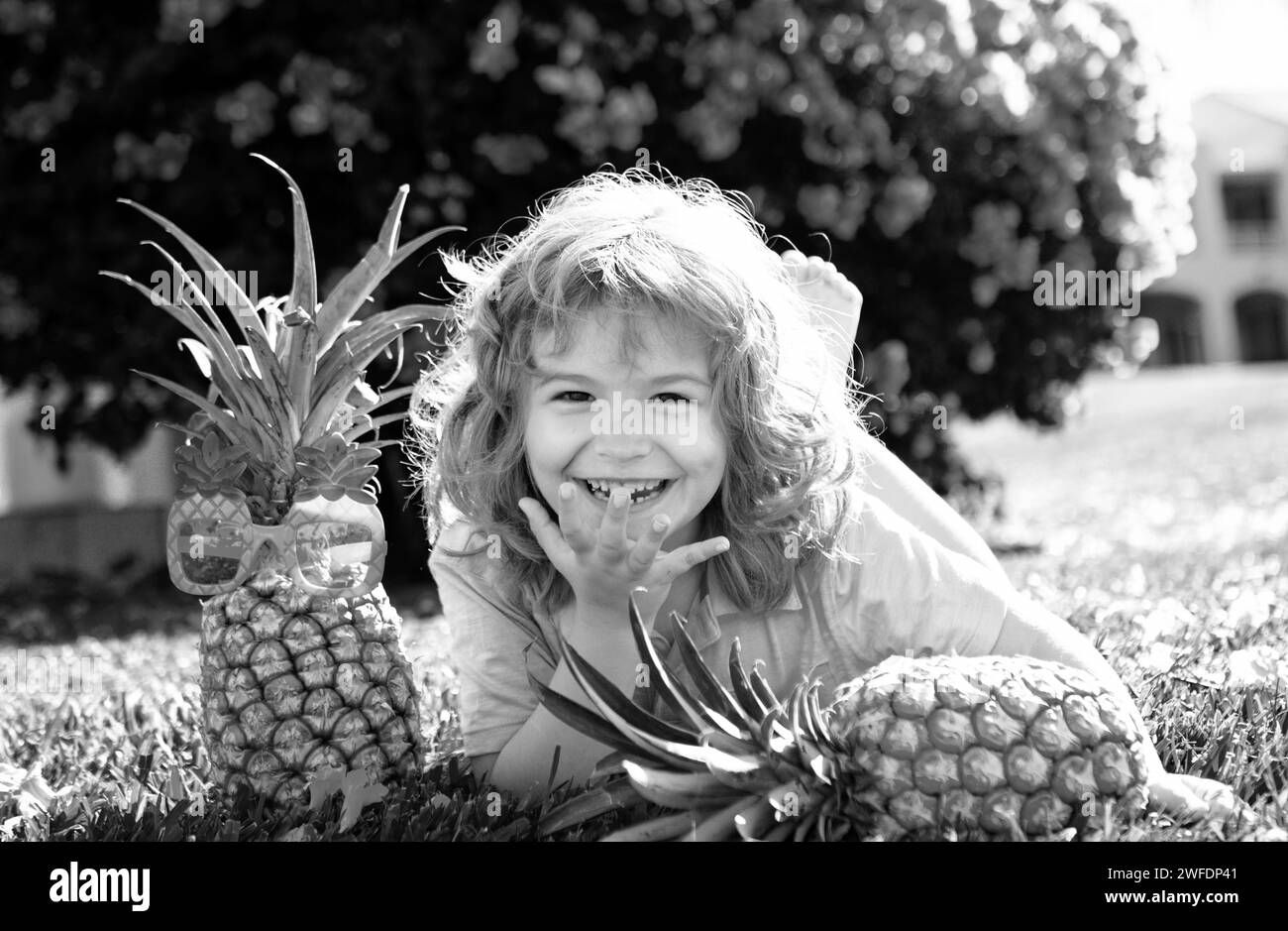 Boy with pineapple on head, plays with fresh tropical fruit outdoorst. Excited funny kid. Child face with amazed expression. Kids summer party. Stock Photo