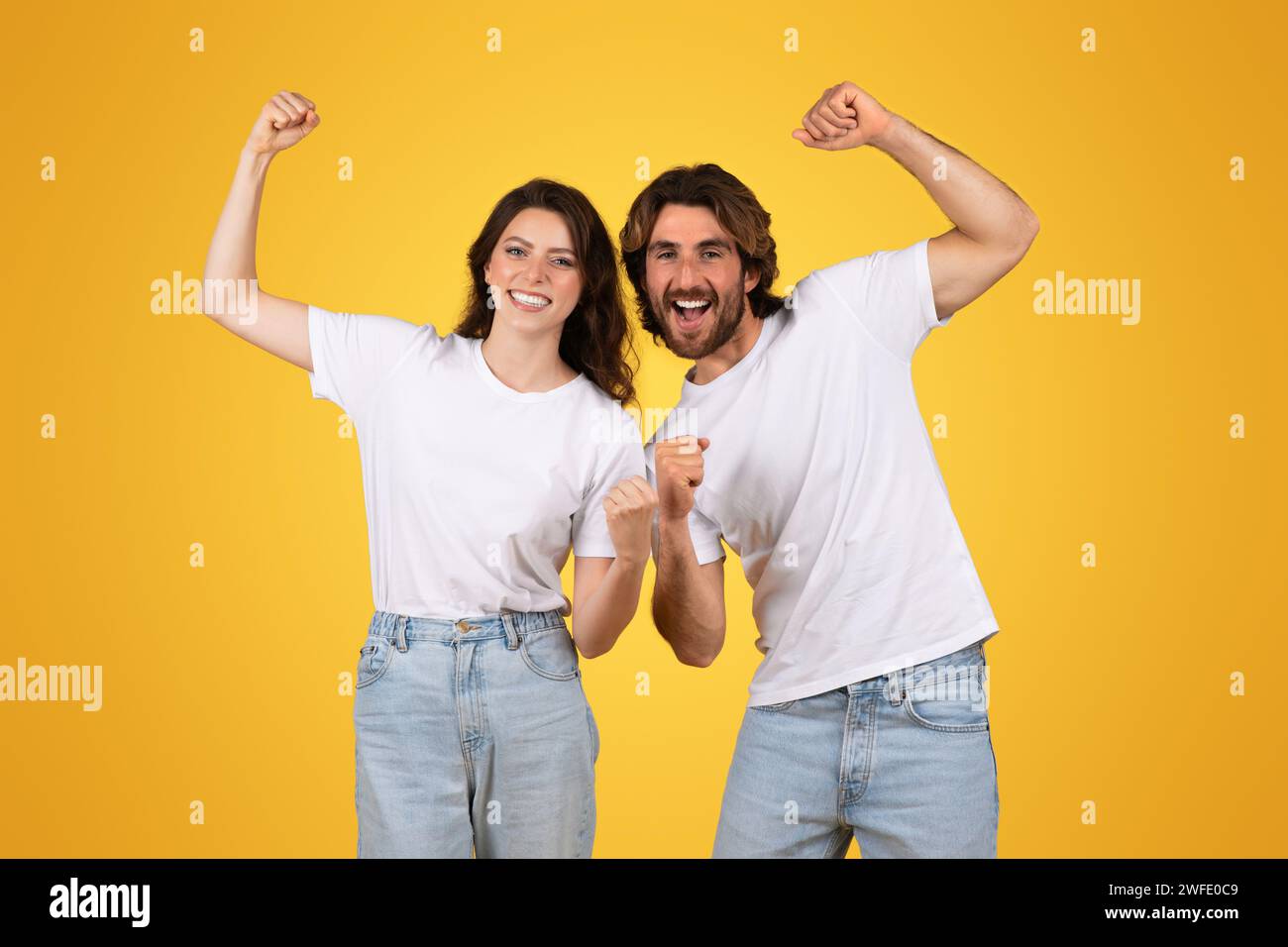 Enthusiastic young couple in white t-shirts and jeans cheerfully raising fists in a victorious gesture Stock Photo