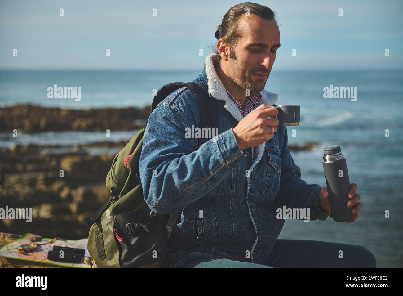 Handsome traveler man holding thermos flask and drinking tea or coffee from stainless steel mug, sitting on the rock by sea, enjoying a happy day outd Stock Photo