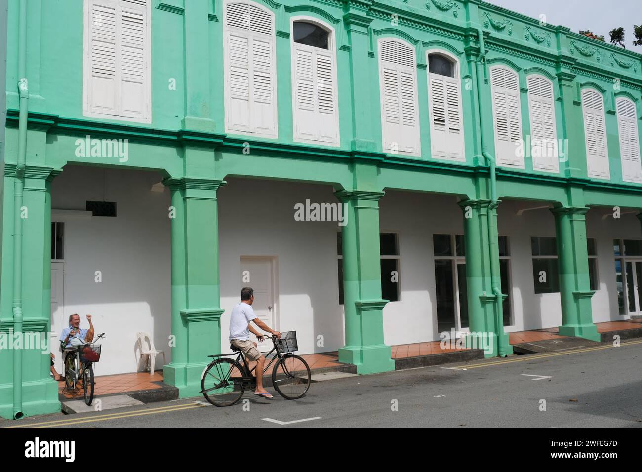 Old Singapore Chinese men on bikes greeting each other by a vibrantly painted shophouse building in Singapore's Little India Stock Photo