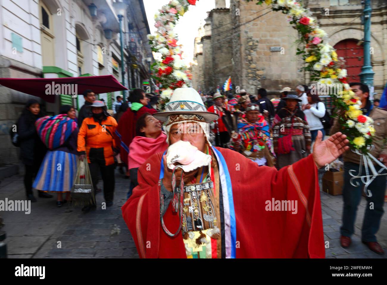 La Paz, BOLIVIA; 24th January 2015. An Aymara shaman blows a conch shell as he walks in front of an ancient illa (or statue) of an Ekeko (an Aymara god of abundance) as it is paraded through the streets of La Paz to celebrate its first appearance at the Alasitas festival, which starts today. The statue is around 2000 years old and was made by the Pucara culture. It was taken from the archaeological site of Tiwanaku to Switzerland in 1858, and returned to Bolivia by the Berne History Museum in November 2014. On the right is part of San Francisco church. Stock Photo