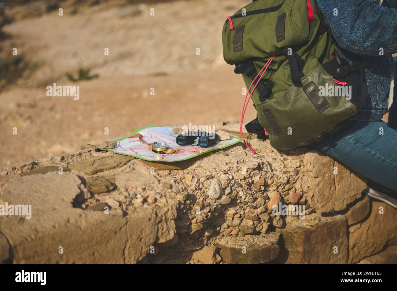 Tourism concept. Close-up binoculars, compass on a map near a backpack of a male tourist sitting on the cliff, enjoying his hiking on the adventure tr Stock Photo