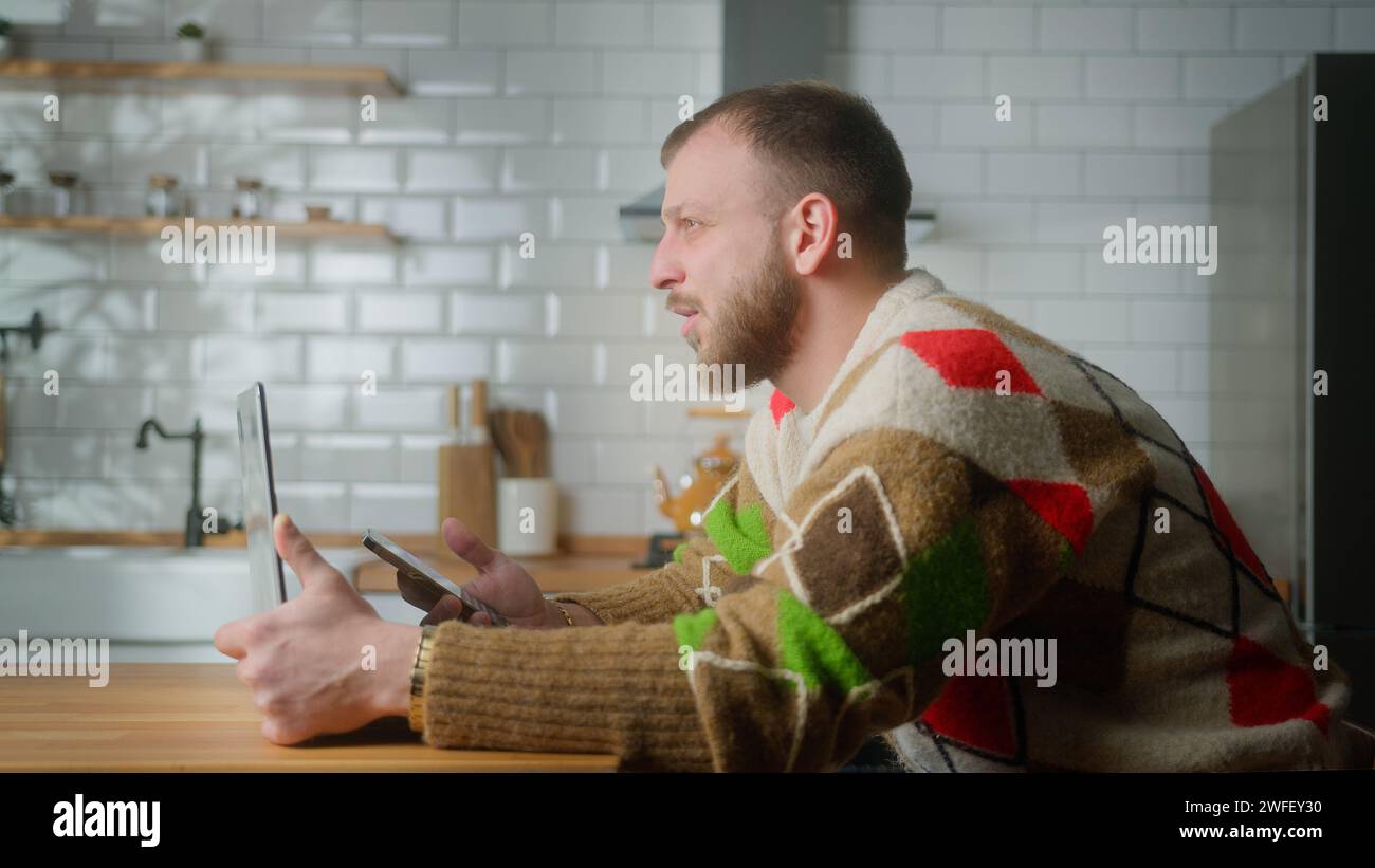 Adult man sit in kitchen with laptop surfing internet on mobile phone, reading media news, scrolling social networks, using mobile applications on sma Stock Photo