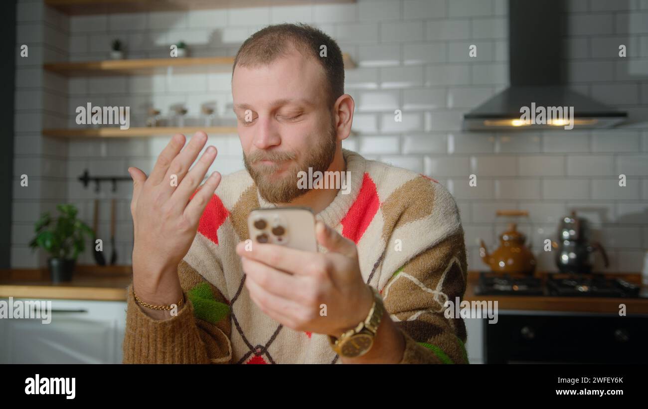 Man sitting in kitchen reacts to what sees, surfing internet on mobile phone, checking email, reading media news, scrolling social medias, using mobil Stock Photo