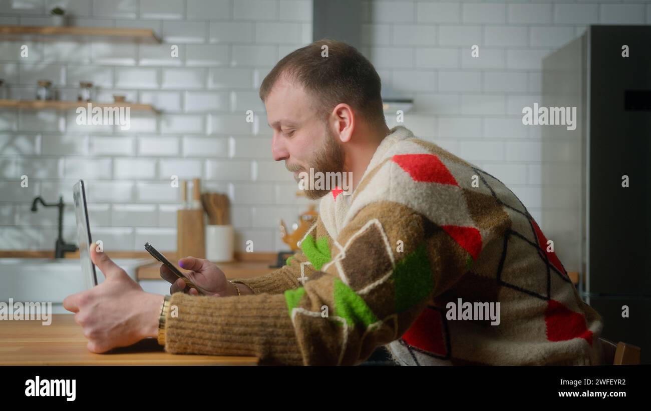 Adult man sit in kitchen with laptop, talking, surfing internet on mobile phone, reading media news, scrolling social networks, using mobile applicati Stock Photo