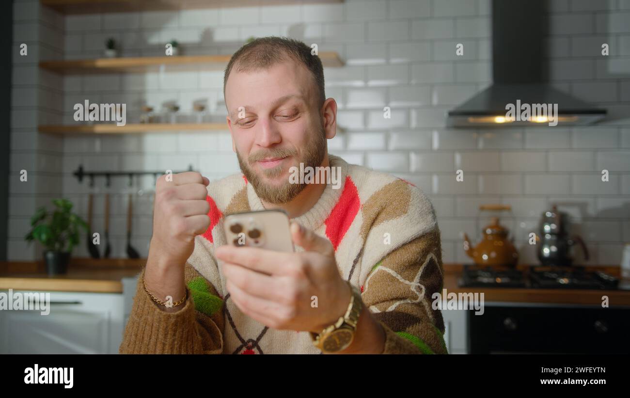 Man sit in kitchen reacts to what sees, surfing internet on mobile phone, checking email, reading media news, scrolling social medias, using mobile ap Stock Photo