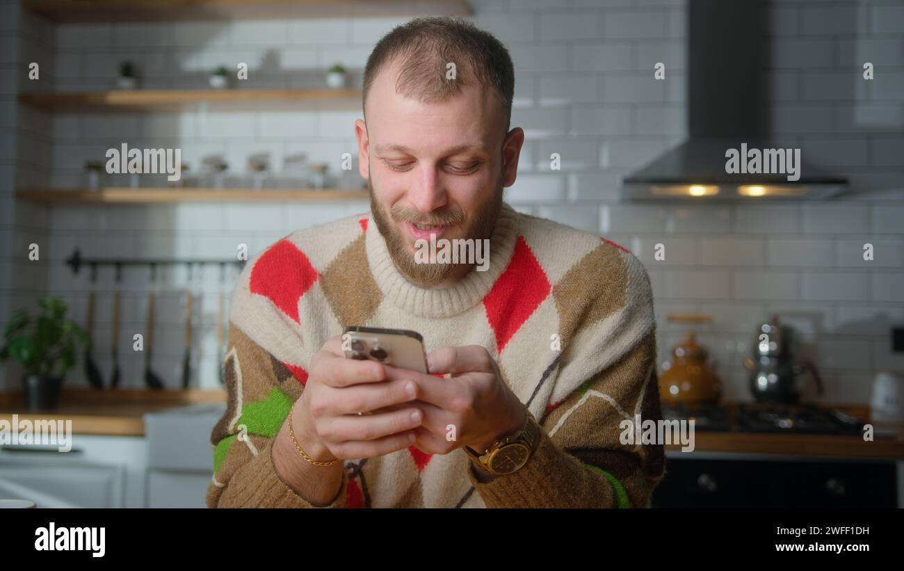 Smiling man sit in kitchen surfing internet on mobile phone, checking email, reading media news, scrolling social networks, using mobile applications Stock Photo