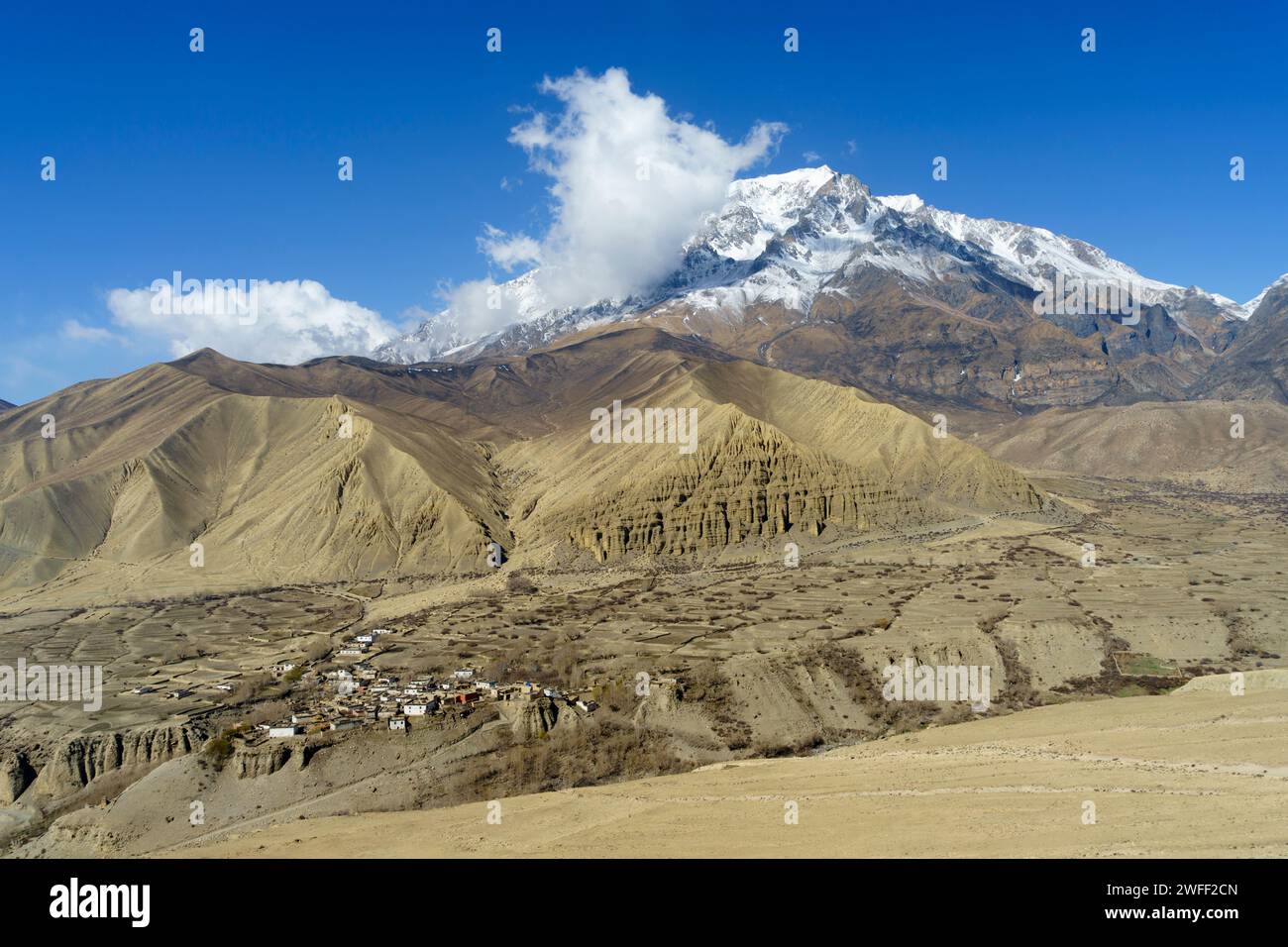Spectacular desertic landscape with snow capped mountains and small village  near Dhakmar,  Upper Mustang region, Nepal. Stock Photo