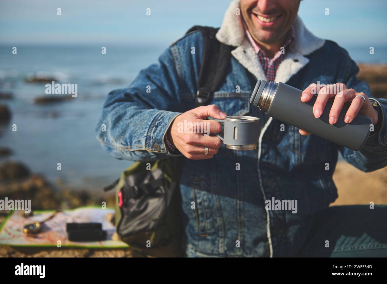 Happy smiling adventurer man in casual denim, pouring hot drink from a thermos flask into a stainless steel mug, sitting on a cliff by sea. Recreation Stock Photo