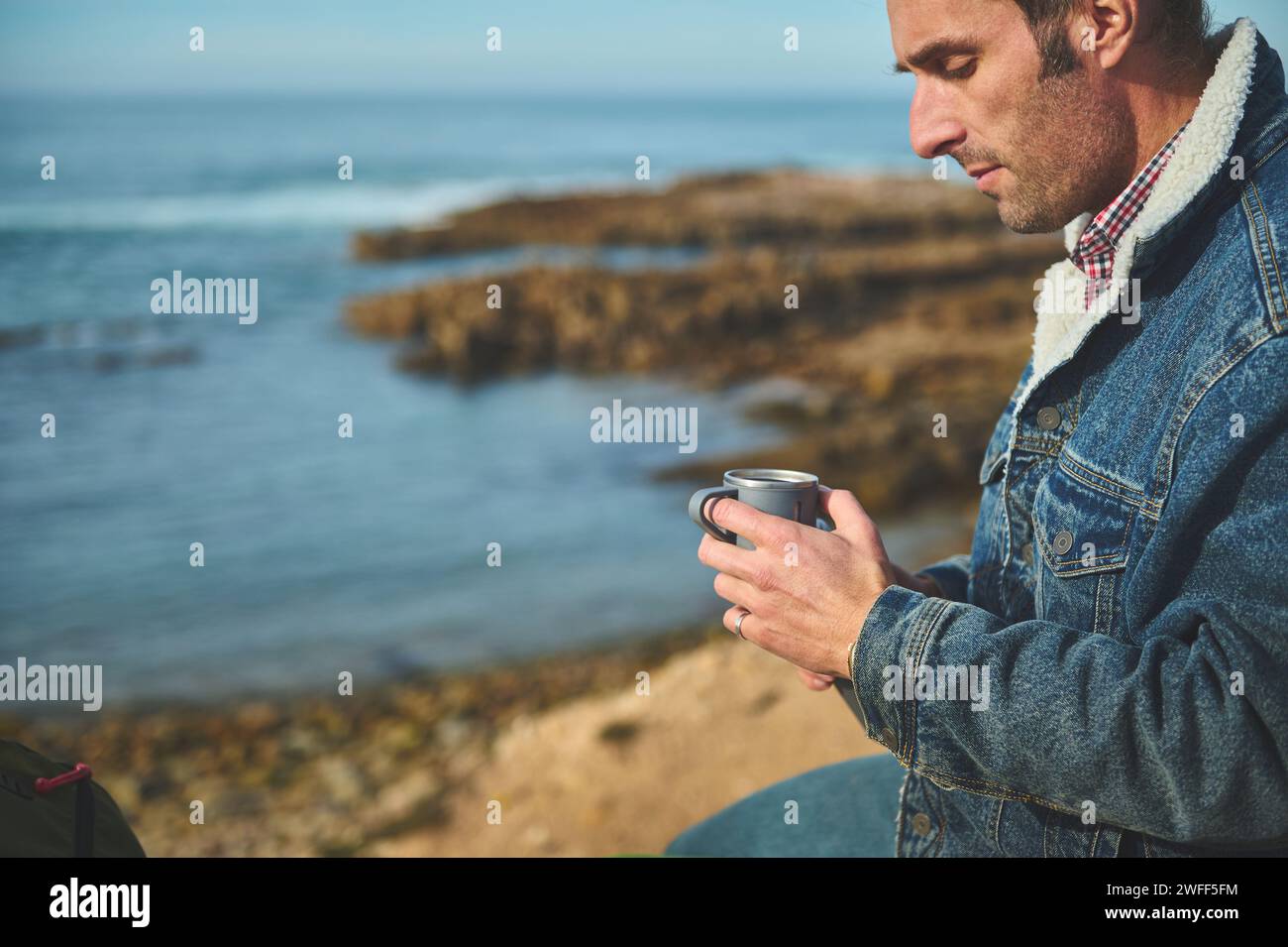 Close-up portrait of Caucasian relaxed serene man traveler holding a mug with hot drink in his hands, relaxing, sitting alone on a rock by sea, enjoyi Stock Photo