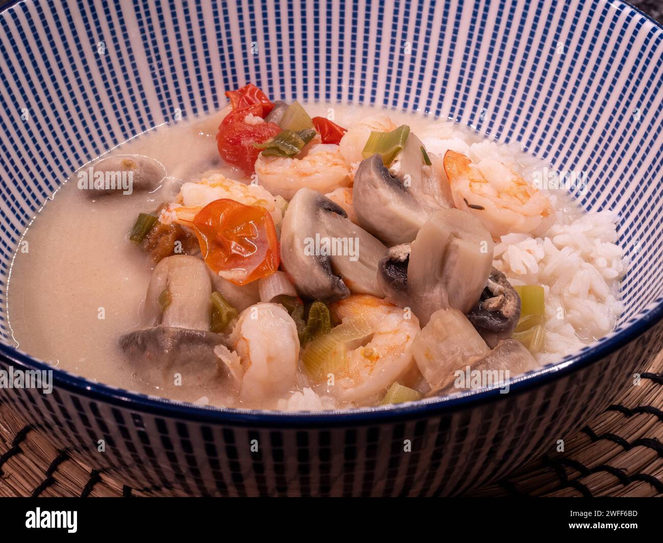 A large blue and white bowl holds a delectable Thai coconut soup infused with spiciness, featuring succulent shrimps, juicy tomatoes, mushrooms, and f Stock Photo