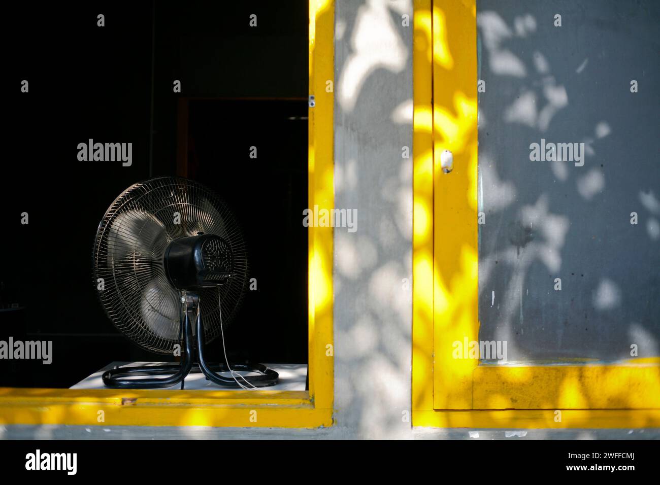 Black electric fan in the yellow window with shadow of leaves in the summer day Stock Photo