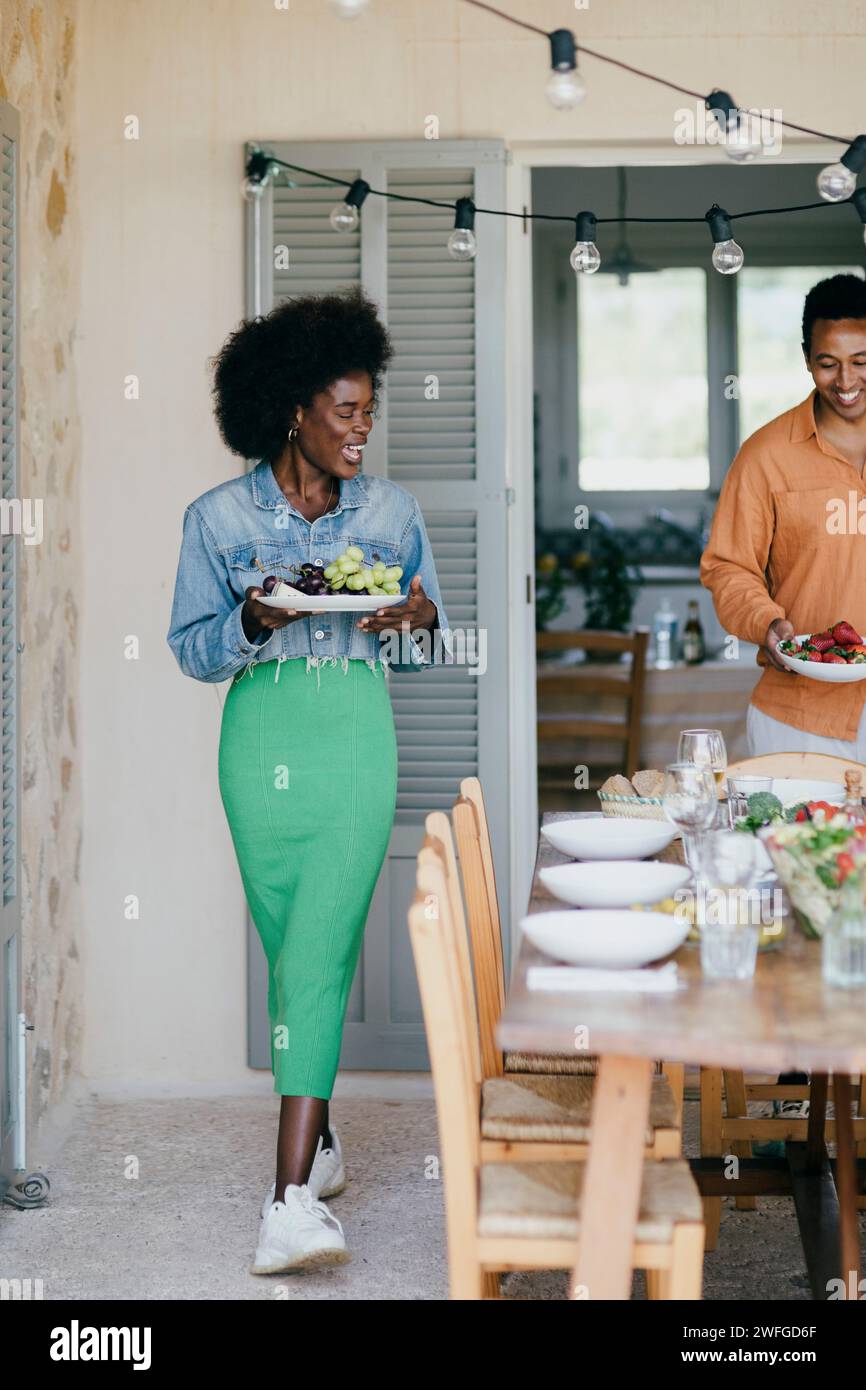 Happy woman with Afro hairstyle walking with plate by dining table in backyard patio Stock Photo