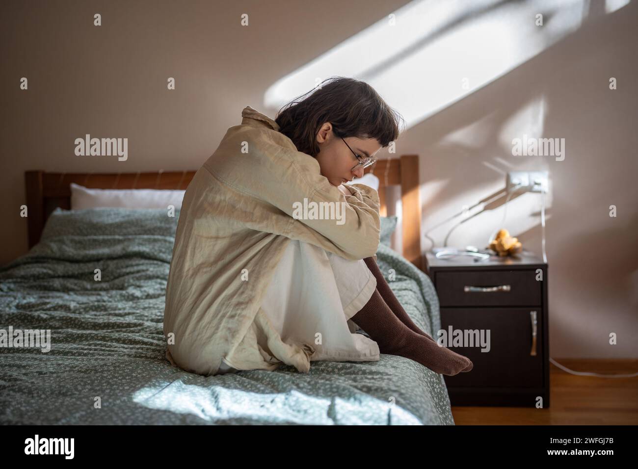 Apathetical teen girl sitting on bed embracing knees feeling procrastination, depression, loneliness Stock Photo