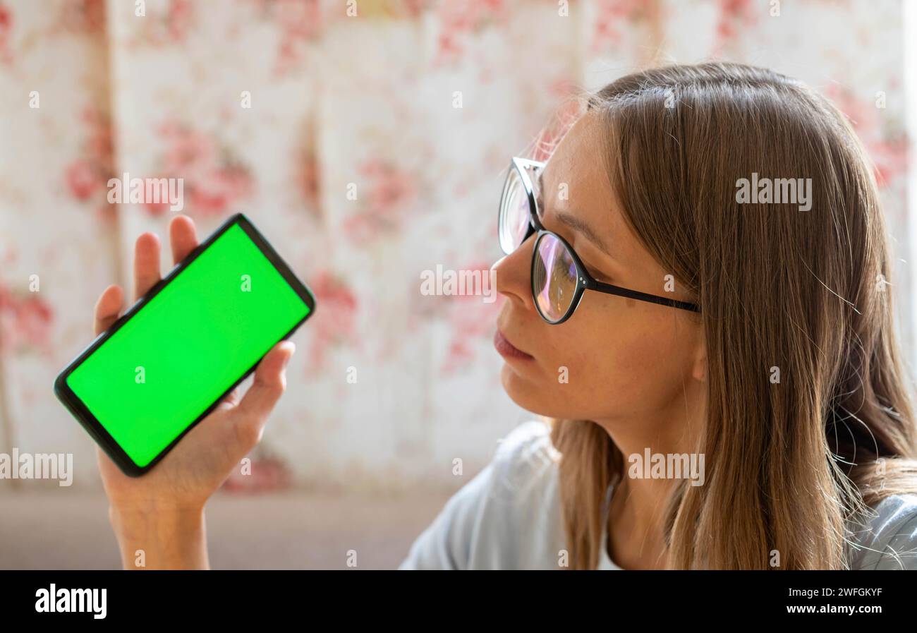 Young girl in romantic home environment holding smartphone with green screen in her hand looking at it. Online pastime, shopping, communication, leisu Stock Photo