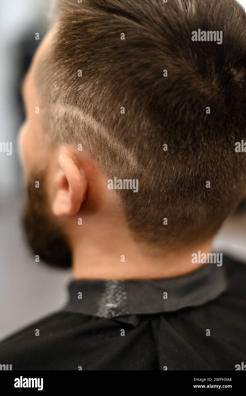 A young man with a beard in a black cape sits on a chair in a barbershop. The client is waiting for a haircut and shaving of the beard. Stock Photo