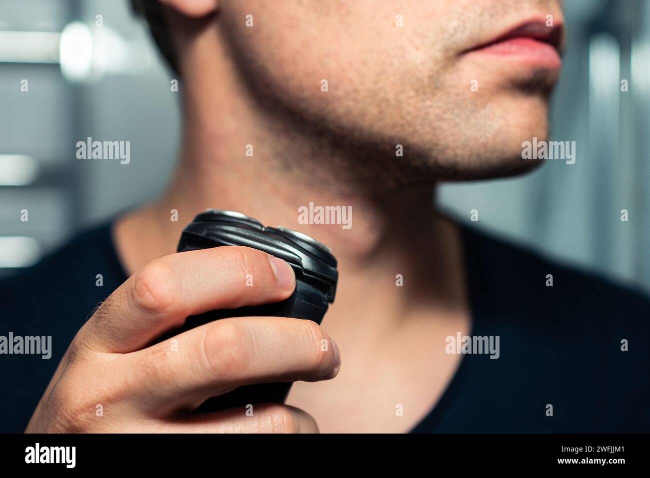 Shaving beard with machine. Man with electric shaver. Trimmer for face hair. Male grooming razor. Mirror in bathroom. Short stubble. Closeup of neck. Stock Photo