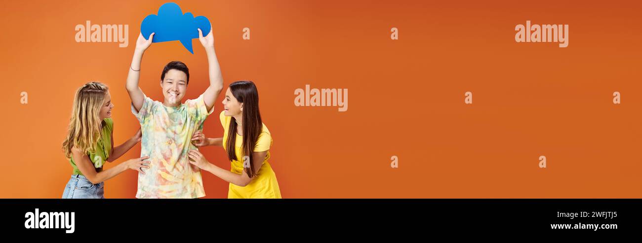 jolly diverse teens posing with blue thought bubble on orange backdrop, friendship day, banner Stock Photo