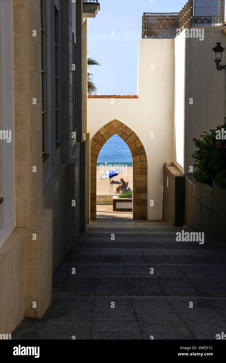 Rota, Cadiz, Spain- October 10, 2023:Tourist sunbathing on Playa de la Costilla beach. Narrow street and arch in the foreground Stock Photo