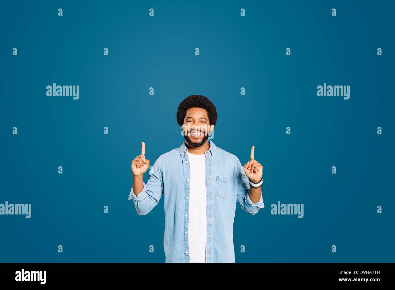 A young man with a warm smile points upwards with both hands, advertising standing against a rich blue backdrop. The gesture and his open expression suggest inspiration or presenting valuable ideas. Stock Photo