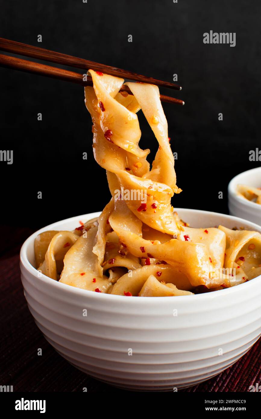 Chopsticks Holding Biang Biang Noodles: Wooden chopsticks lifting belt noodles topped with hot and spicy chili oil with pork mince out of a bowl Stock Photo