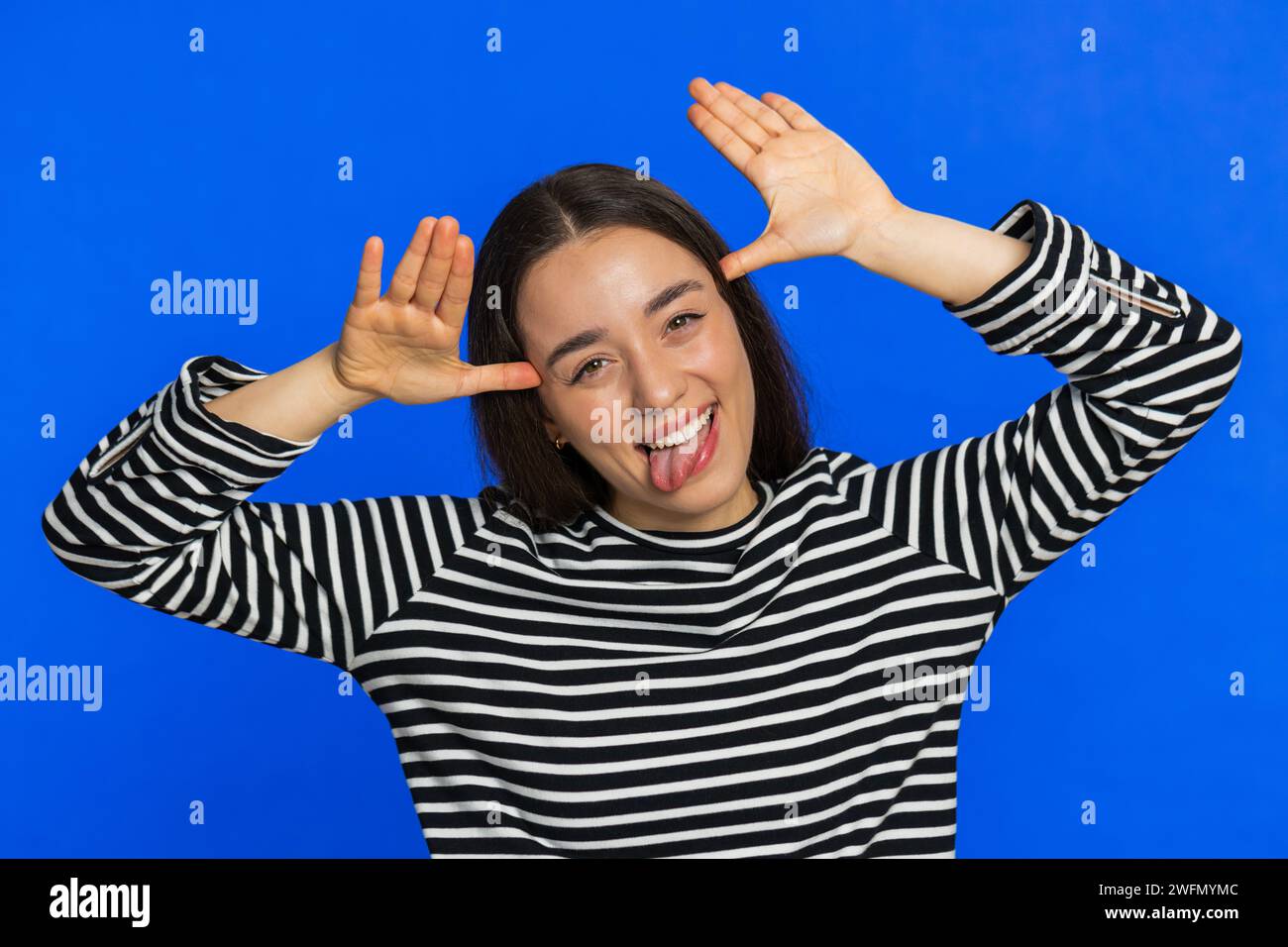 Funny Caucasian woman showing tongue making faces at camera, fooling around, joking, aping with silly face, teasing, bullying, abuse disrespect. Joyful young girl isolated on blue background indoors Stock Photo