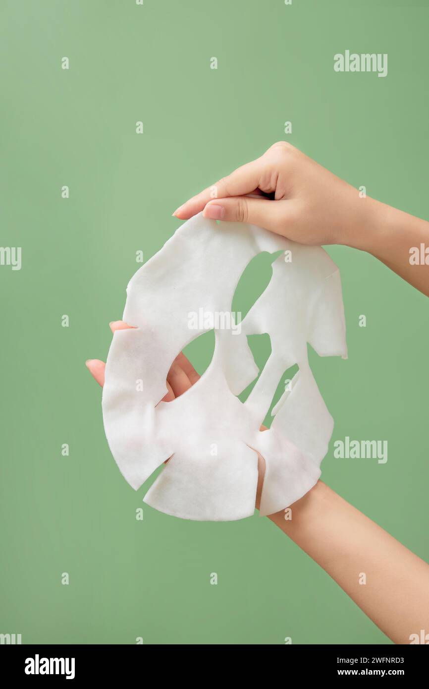 A white skincare mask is held on hand model against a light green background. Skincare mask treatment for face. Beauty product Stock Photo