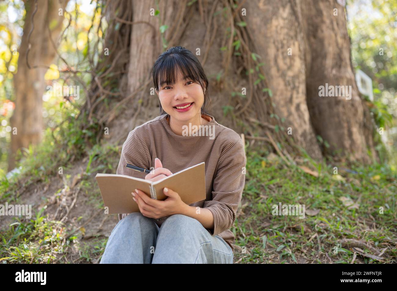 A charming young Asian woman is reading book and finding her inspiration while resting under a tree in a park. leisure time, lifestyle Stock Photo