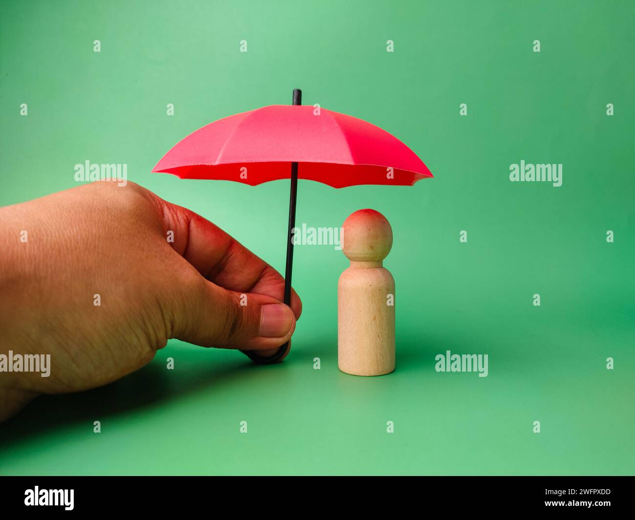 Someone hand holding red umbrella to protect wooden peg doll on a green background. Stock Photo